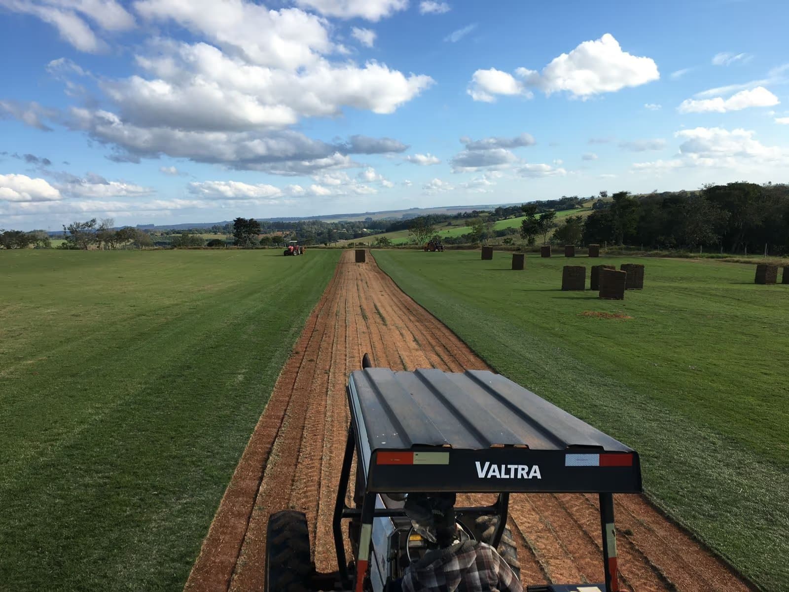 Trator colhendo uma faixa de grama num campo de cultivo, com a terra vermelha exposta e pilhas de placas ao fundo